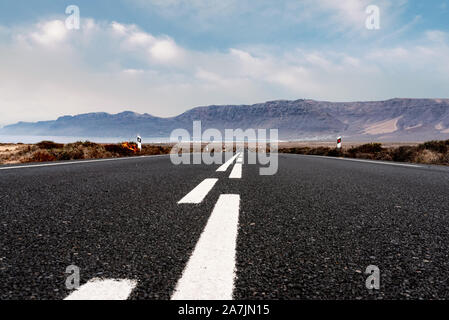 Lungo rettilineo vuoto strada asfaltata attraverso il paesaggio arido contro l'oceano e la gamma della montagna a Lanzarote isole Canarie Foto Stock