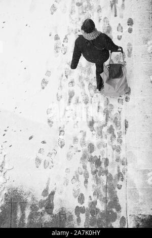 Sfocata donna camminare durante il traino di un carrello di shopping bag durante la nevicata, elevato angolo vista da dietro in bianco e nero Foto Stock