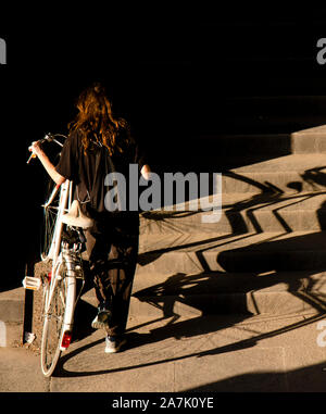 Una ragazza adolescente in nero spingendo la bici bianco fino al pubblico le scale di luce e ombre, da dietro Foto Stock