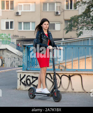 A Belgrado, in Serbia - Ottobre 25, 2019: Una ragazza adolescente in abito rosso e giacca di pelle in sella ad una scooter elettrico sulla strada di città marciapiede Foto Stock