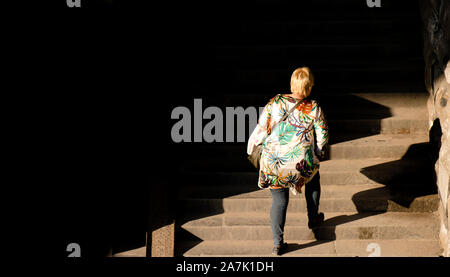 Una bionda Taglie donna a piedi fino al pubblico le scale di luce e ombre, da dietro Foto Stock