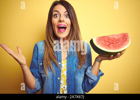 Giovane bella donna mangiare fresco e salutare fetta di anguria su sfondo giallo molto felici ed eccitati, vincitore espressione celebrando la vittoria screa Foto Stock