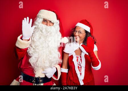 La mezza età giovane indossando Santa costume e vetri isolati su sfondo rosso rinuncia a dire ciao felice e sorridente, gentile gesto di benvenuto Foto Stock