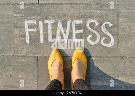 La parola scritta fitness sul marciapiede grigio con gambe di donna, vista da sopra Foto Stock