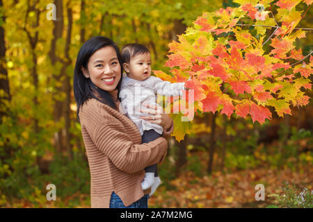 Asian madre cinese holding carino adorabile bambina sulle mani in autunno autunno park outdoor con giallo arancione foglie degli alberi. Halloween o ringraziamento Foto Stock