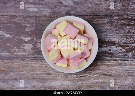 Rosa e giallo marshmallows in una ciotola bianco nel mezzo di uno sfondo di legno, vista dall'alto Foto Stock