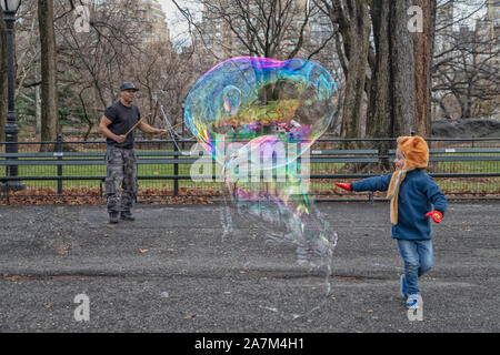 New York, STATI UNITI D'AMERICA-dicembre 17,2018: un uomo soffia bolle grandi nel Central Park di New York con un bellissimo bambino contento di toccarlo prima che rompe il giorno Foto Stock