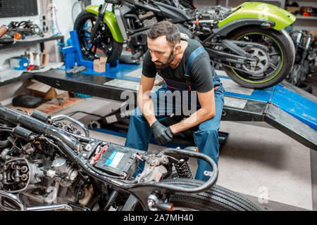 Ritratto di un bel barbuto biker in tute da lavoro seduta vicino alla moto durante la riparazione in officina Foto Stock