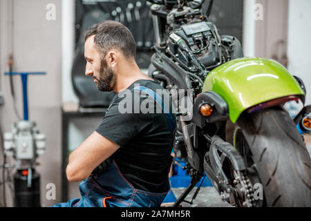 Ritratto di un bel barbuto biker in tute da lavoro seduta vicino alla moto durante la riparazione in officina Foto Stock
