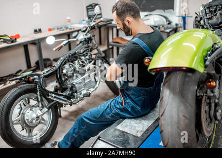 Ritratto di un bel barbuto biker in tute da lavoro seduta vicino alla moto durante la riparazione in officina Foto Stock