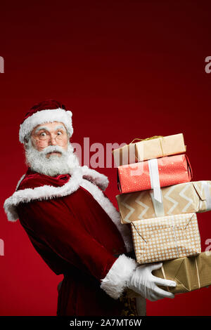 Vita ritratto della favola di Santa holding pila di regali di Natale su sfondo rosso, spazio di copia Foto Stock