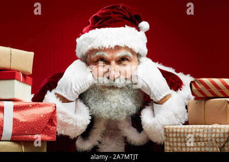 Close up ritratto della favola di Santa guardando la fotocamera mentre posa con i regali di Natale su sfondo rosso Foto Stock
