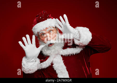 Vita ritratto del classico Babbo Natale facendo facce buffe mentre posa contro lo sfondo di colore rosso in studio, spazio di copia Foto Stock