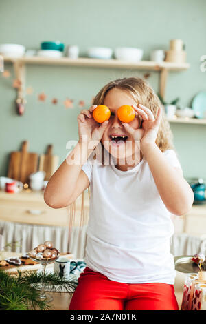 Ritratto di un allegro bambina con i tangerini nei suoi occhi, natale, cucina, casa accogliente Foto Stock