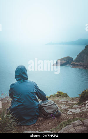 Vista posteriore di un escursionista donna seduta sul bordo di una scogliera con un impermeabile blu e zainetto guardando l'orizzonte e il mare. Foto Stock