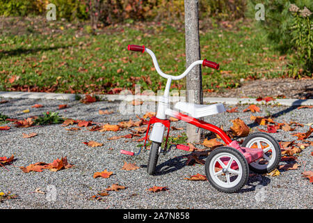 Un rosso e bianco il triciclo è parcheggiato all'esterno in un parco sulla ghiaia. Il trike è circondato da foglie cadute, mostrando la stagione autunnale. Foto Stock