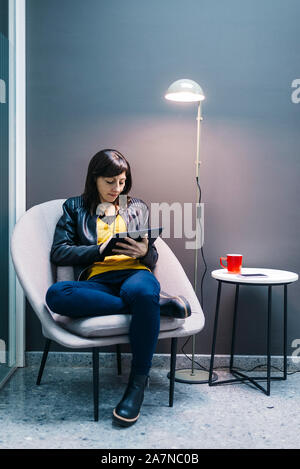 La donna a casa seduti sulla sedia moderna e utilizzando computer tablet, rilassante in ufficio Foto Stock