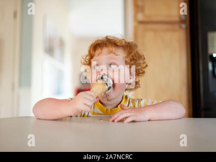 Confuso toddler boy mangia gelato mentre è seduto al banco in cucina Foto Stock