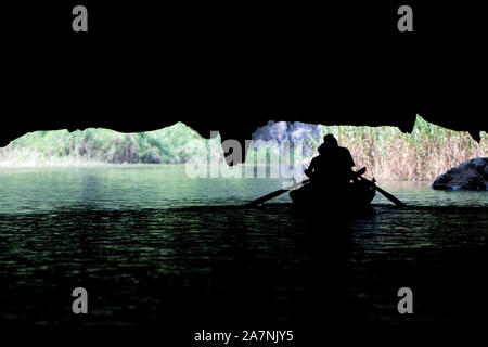 Una riga barca piena di turisti su una crociera sul fiume tour emerge da sotto una grande grotta di calcare in Tam Coc, nel Vietnam del Nord Foto Stock