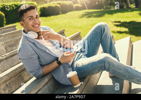 Per il tempo libero all'aria aperta. Elegante ragazzo indossa le cuffie guardare seduti sulle scale nel parco con tazza di caffè rilassato sorridente Foto Stock