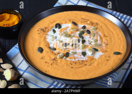 Close-up di cremosa butternut arrosto zuppa di zucca cosparso con i semi di zucca in una ciotola nero su un tavolo di legno, ingredienti in background su un s Foto Stock