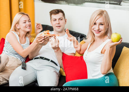 Junk food o cibo sano. Attraente sportivo caucasica donna bionda mangiare apple mentre i suoi amici gustando la pizza, facendo segno di stop, sorridente guardando Foto Stock