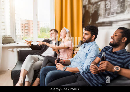 Felice multiculturale giovani amici sono in appoggio a casa seduti sul divano con bevande analcoliche, sorridente la visione di gioco di sport in TV. Home partito, incontri sportivi Foto Stock