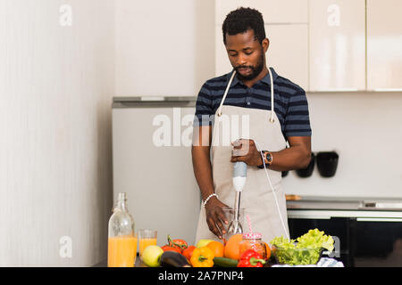 Giovani barbuto americano africano uomo che indossa polo t-shirt grembiule e la preparazione di un alimento sano, con blender per sminuzzare gli ingredienti, rendendo smo vegetale Foto Stock