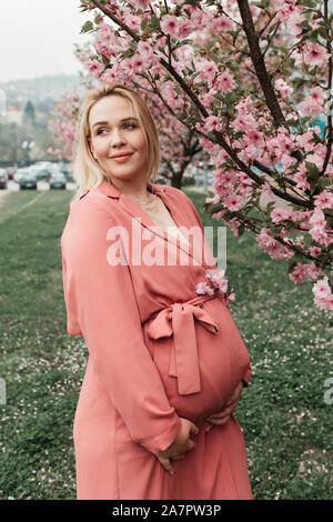 Belle Donne incinte in abito rosa fotografata accanto ad alberi fioriscono in primavera. Foto Stock