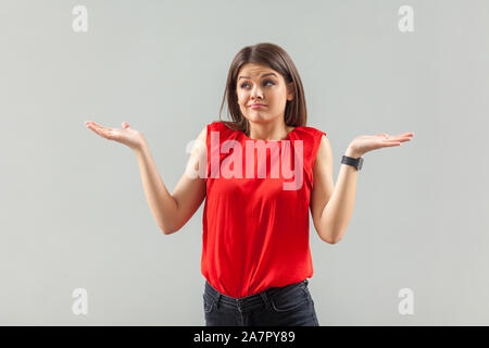 Io non sono certo! Ritratto di confuso bella bruna giovane donna in camicia rossa in piedi con le braccia sollevate, guardando lontano e fare'nt sanno cosa fare. ind Foto Stock