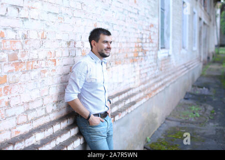 Giovane bello guy standing vicino al muro davanti a sé sembra, sorride e tiene le mani nelle tasche dei pantaloni. Foto Stock