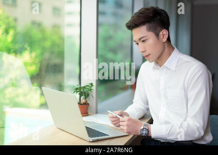 Un occupato sorridente giovane imprenditore con corti capelli neri e un bianco button down shirt lavora davanti ad un laptop a una scrivania in un ufficio Foto Stock