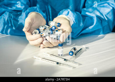 Dottore in un blu camici chirurgici e maschera e tiene in mano le bottiglie con insulina per il diabete. Molte le siringhe e farmaci per iniezione sul tavolo bianco Foto Stock