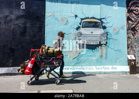 Berlino muro graffiti Migrant senza casa o turista economico con supermercato Tram che passa intorno Trabant auto a East Side Gallery Germania Foto Stock