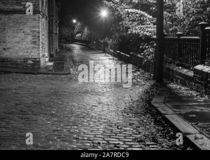 La lampada accesa strade acciottolate di Saltaire, raffigurato in moody Black & White, rivelano mentre questa strada è spesso utilizzato da aziende di pellicola per scatti di posizione Foto Stock