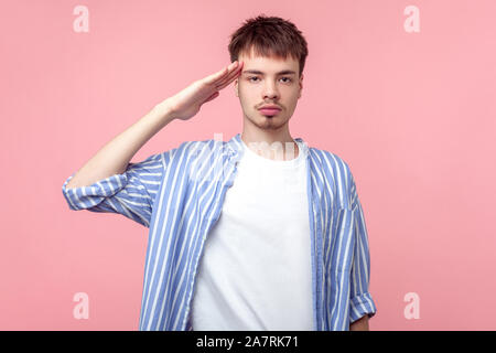 Yes Sir! Ritratto di giovane diligente i capelli castani uomo con piccola barba e baffi in casual striped shirt cercando grave alla fotocamera e salutando ind. Foto Stock