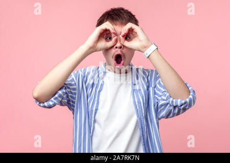 Ritratto di stupito curiosi i capelli castani uomo con piccola barba e baffi in camicia casual guardando attraverso le dita gesticolando binocolo, scioccato ga Foto Stock