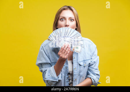 Ritratto di sorpreso giovane donna in denim shirt che spuntavano di dollari ventola, nascondendo la sua metà faccia e guardare la fotocamera con grandi occhi attoniti. interna s Foto Stock