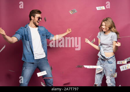 Ritratto di un felice elegantemente vestito giovane celebrando in piedi sotto la doccia di denaro isolate su sfondo rosa. Foto Stock