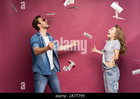 Ritratto di un felice elegantemente vestito giovane celebrando in piedi sotto la doccia di denaro isolate su sfondo rosa. Foto Stock