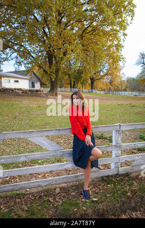 Una bella ragazza in un maglione rosso sorge nei pressi di un bianco staccionata in legno. Bella autunno caldo Foto Stock