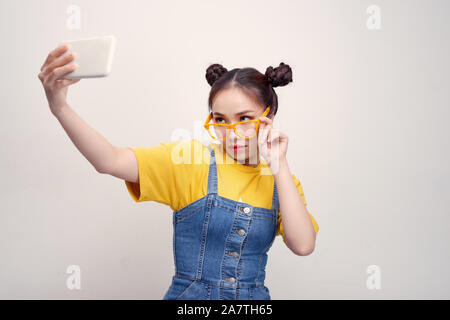 Giovane bella ragazza asiatica che indossa un paio di jeans dungaree e giallo bicchieri, tenendo selfie, sorridente Foto Stock