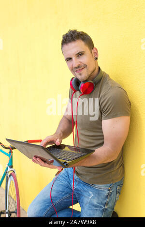 Giovane uomo che lavora con il computer portatile appoggiato contro una parete gialla con la sua fixie accanto a lui Foto Stock