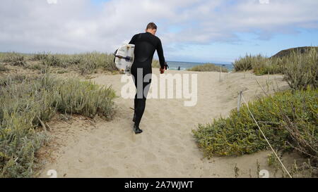 Un uomo solitario che indossa una muta nera e portante una tavola da surf è camminare su un sentiero di sabbia sulle dune verso l'Oceano Pacifico e nuvoloso cielo blu Foto Stock