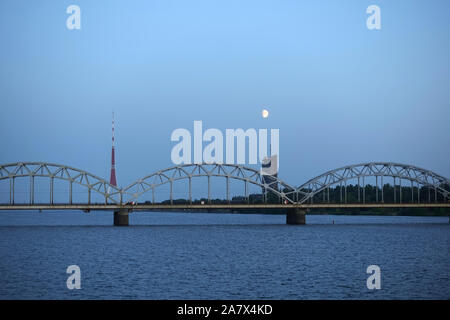 Metallo lungo la ferrovia ponte ad arco attraverso il fiume Daugava a Riga contro stand alone edificio, la torre della TV e luminosa luna vista in avanti sul Cielo di tramonto Foto Stock