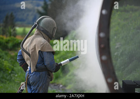 Un uomo pieno weating ingranaggio di sicurezza utilizza la massa di vetro per sabbiatura l'involucro di acciaio di carne di una macchina essiccatrice per una sardigna Foto Stock