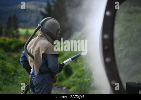 Un uomo weating piena sicurezza sandblasts marcia l'involucro di acciaio di carne di una macchina essiccatrice per una sardigna con vetro macinato Foto Stock