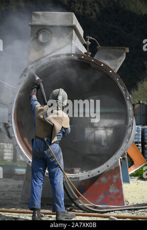 Un uomo pieno weating ingranaggio di sicurezza utilizza la massa di vetro per sabbiatura l'involucro di acciaio di carne di una macchina essiccatrice per una sardigna Foto Stock