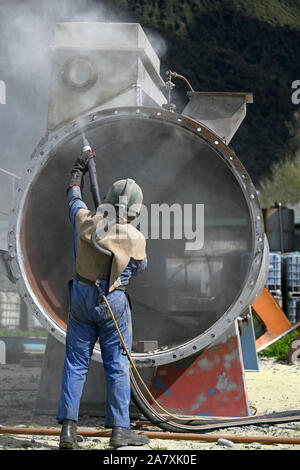 Un uomo pieno weating ingranaggio di sicurezza utilizza la massa di vetro per sabbiatura l'involucro di acciaio di carne di una macchina essiccatrice per una sardigna Foto Stock