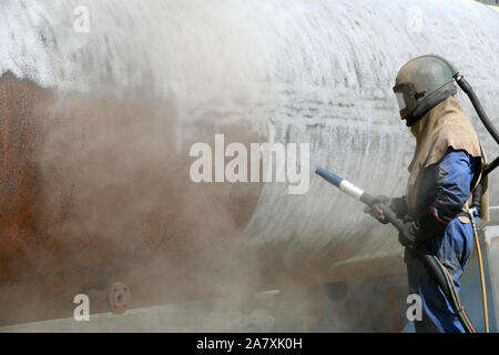 Un uomo weating piena sicurezza sandblasts marcia l'involucro di acciaio di carne di una macchina essiccatrice per una sardigna con vetro macinato Foto Stock